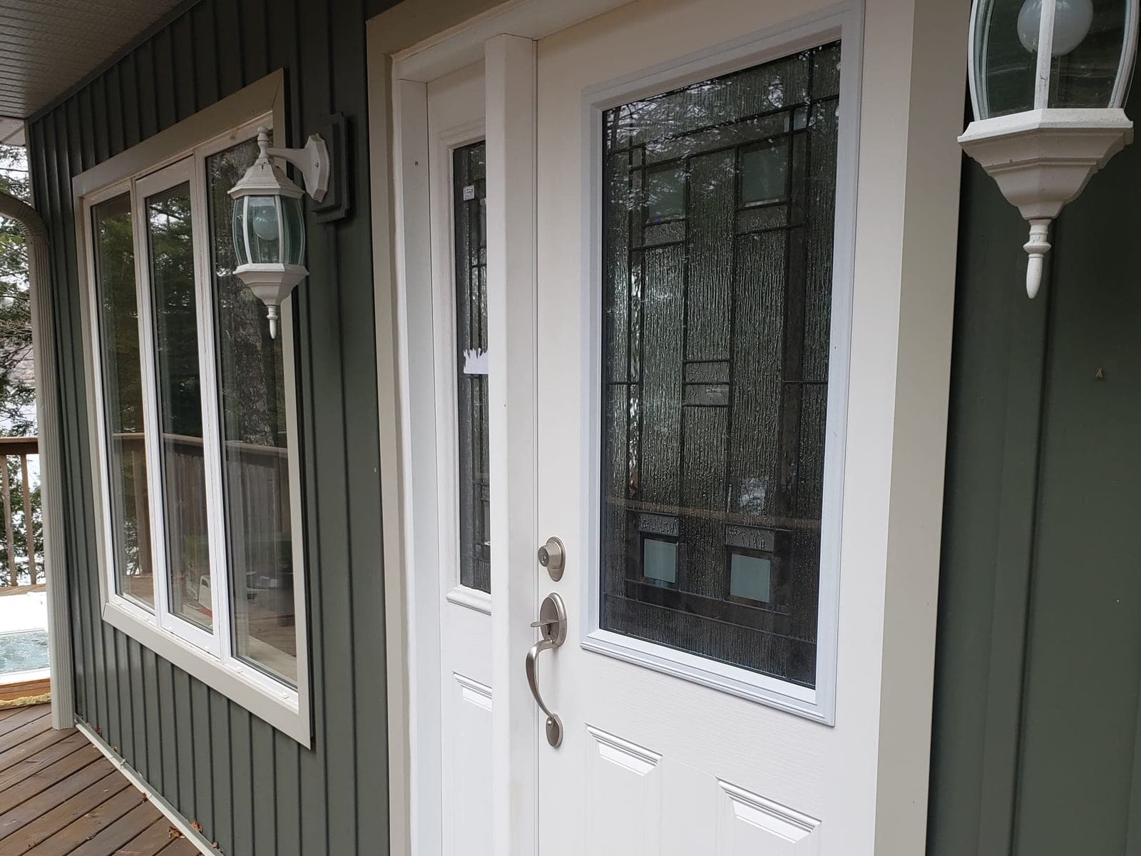 White entry door with a leaded-glass insert and sidelite beside a triple casement window in dark green board-and-batten siding