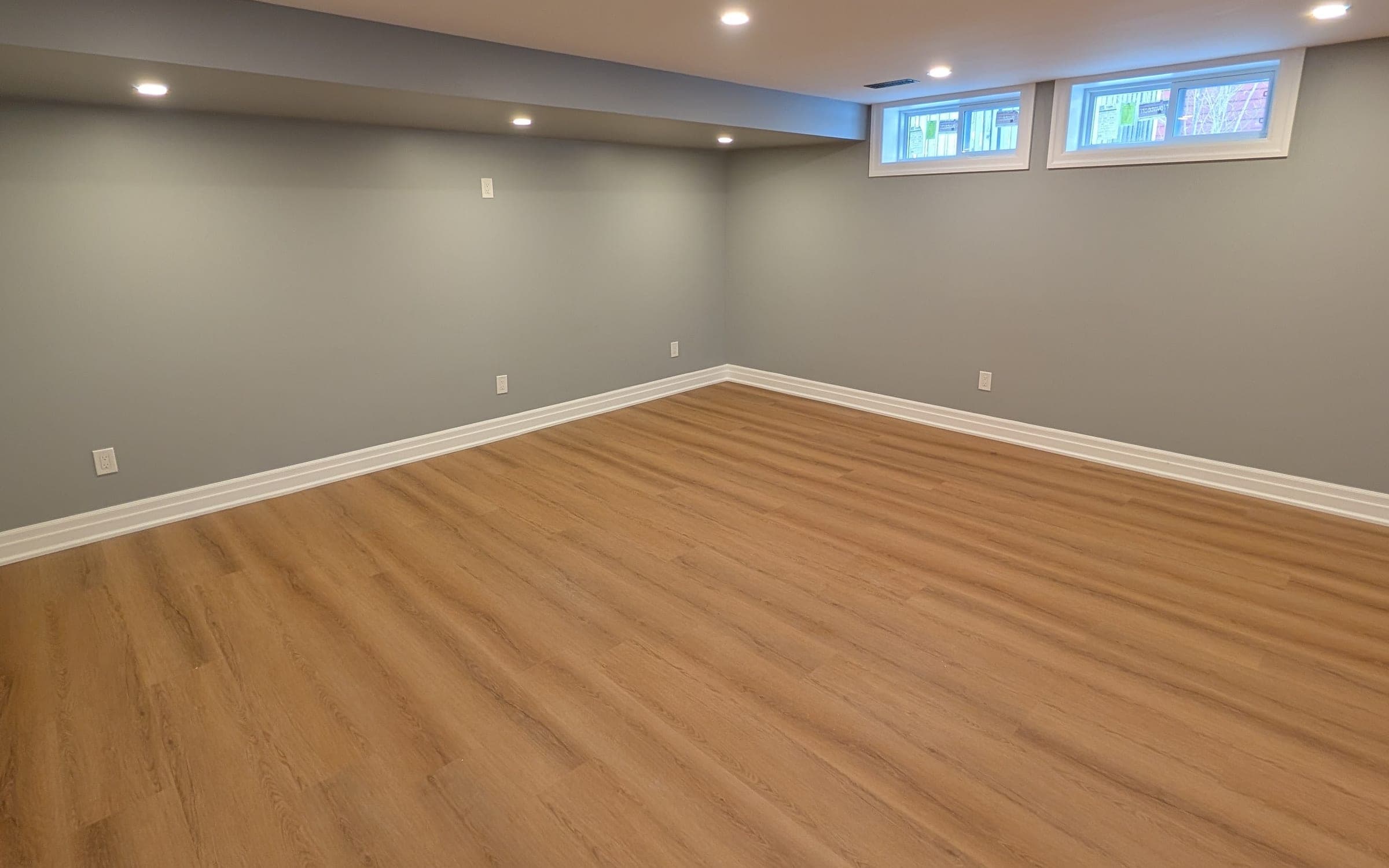 The same basement finished with grey painted walls, white baseboards, light oak vinyl plank flooring, recessed ceiling lights, and two trimmed windows