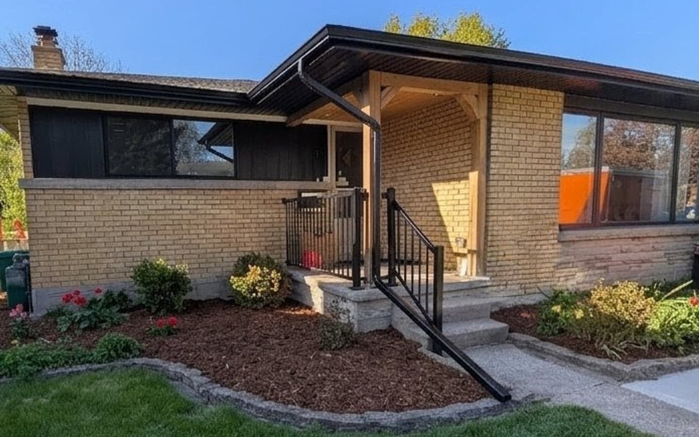The same bungalow after renovation, with a new timber post-and-beam entry canopy, black metal railings, stone-faced front steps, mulched garden beds with stone edging, and fresh sod