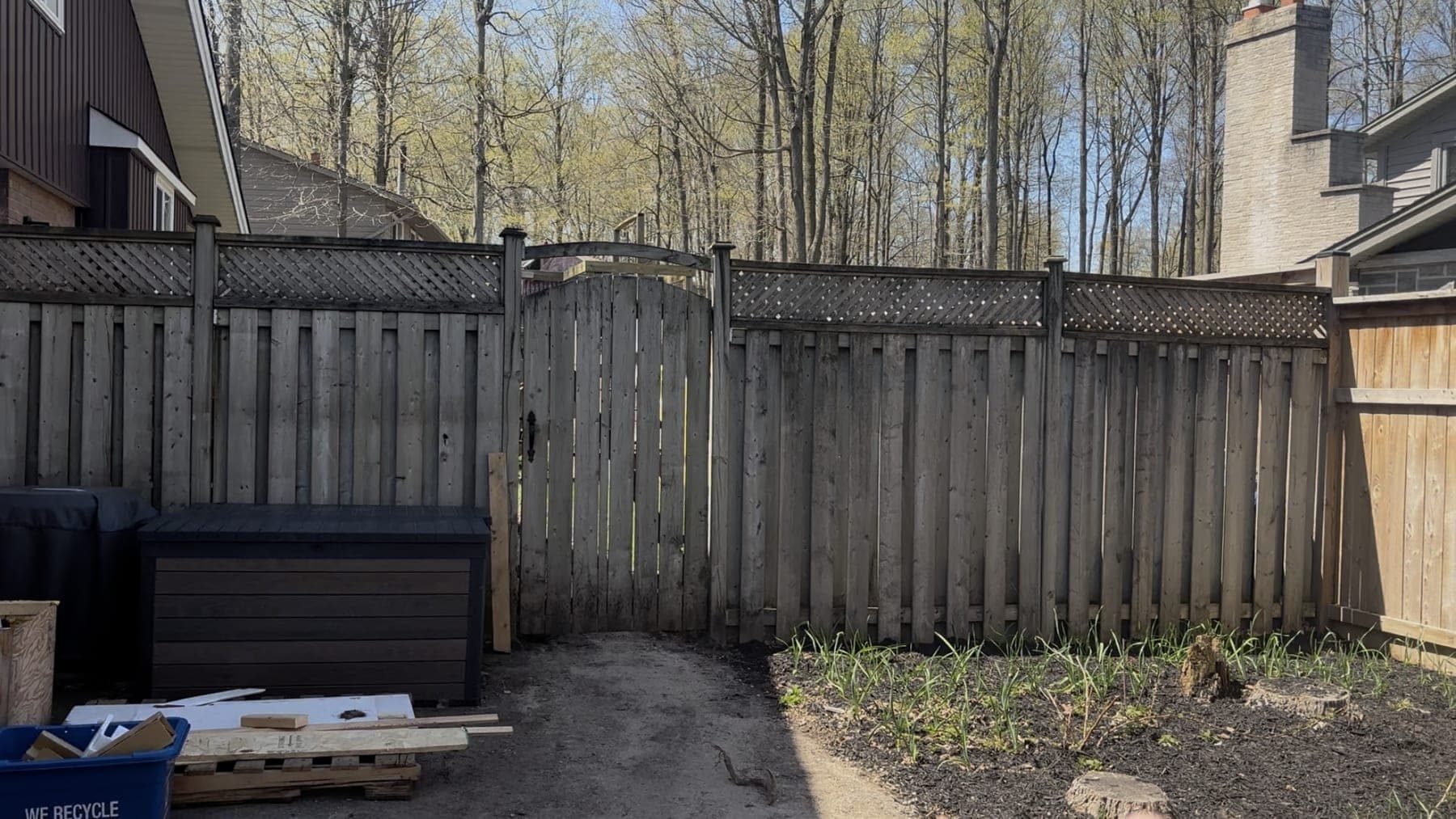 Weathered grey board fence with a lattice topper and a sagging arched gate, beside a bare dirt side yard holding a deck box, recycling bin, and stacked offcuts
