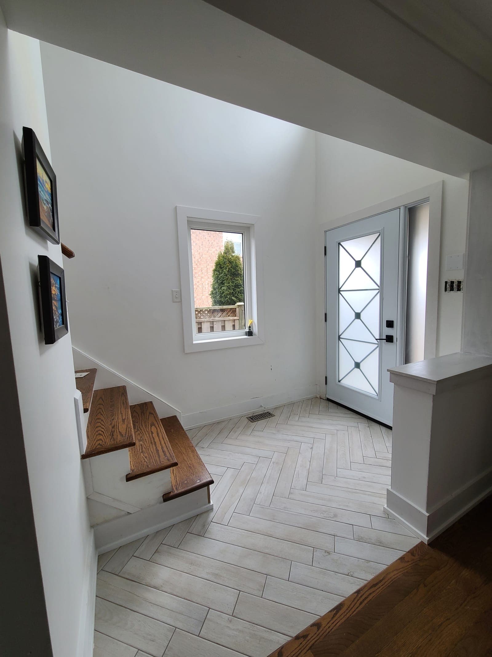Entry foyer with pale herringbone tile flooring, oak stair treads, and a white front door with geometric glass